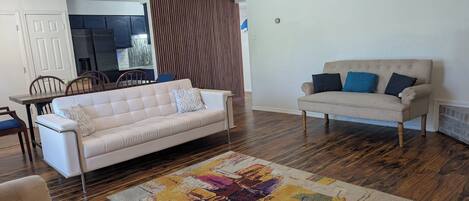 A living room with a white sofa, a beige sofa, a patterned rug, wooden flooring, a ceiling fan, and a kitchen in the background.