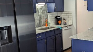 A modern kitchen with dark blue cabinets, a stainless steel refrigerator, a coffee maker, and a trash can.