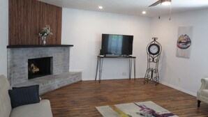 A living room with a fireplace, a flat-screen TV mounted on a stand, a decorative clock, and a colorful rug.