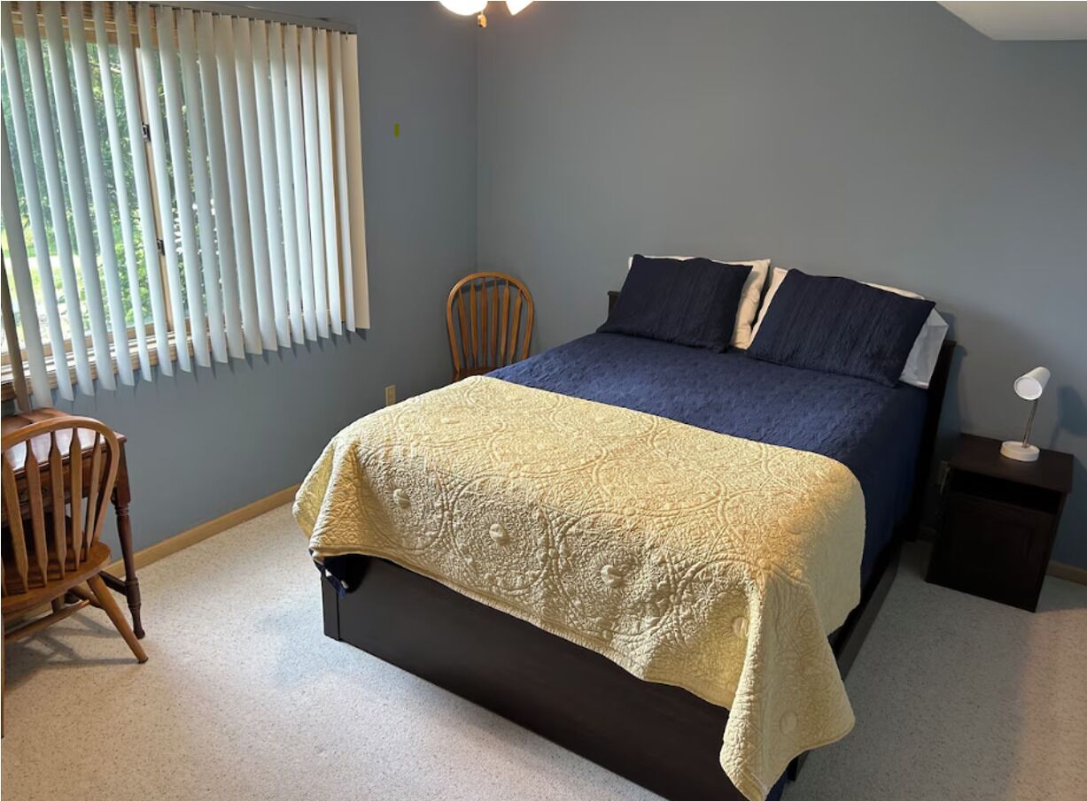 A bedroom with a bed, a chair, a bedside table, and a window with blinds.