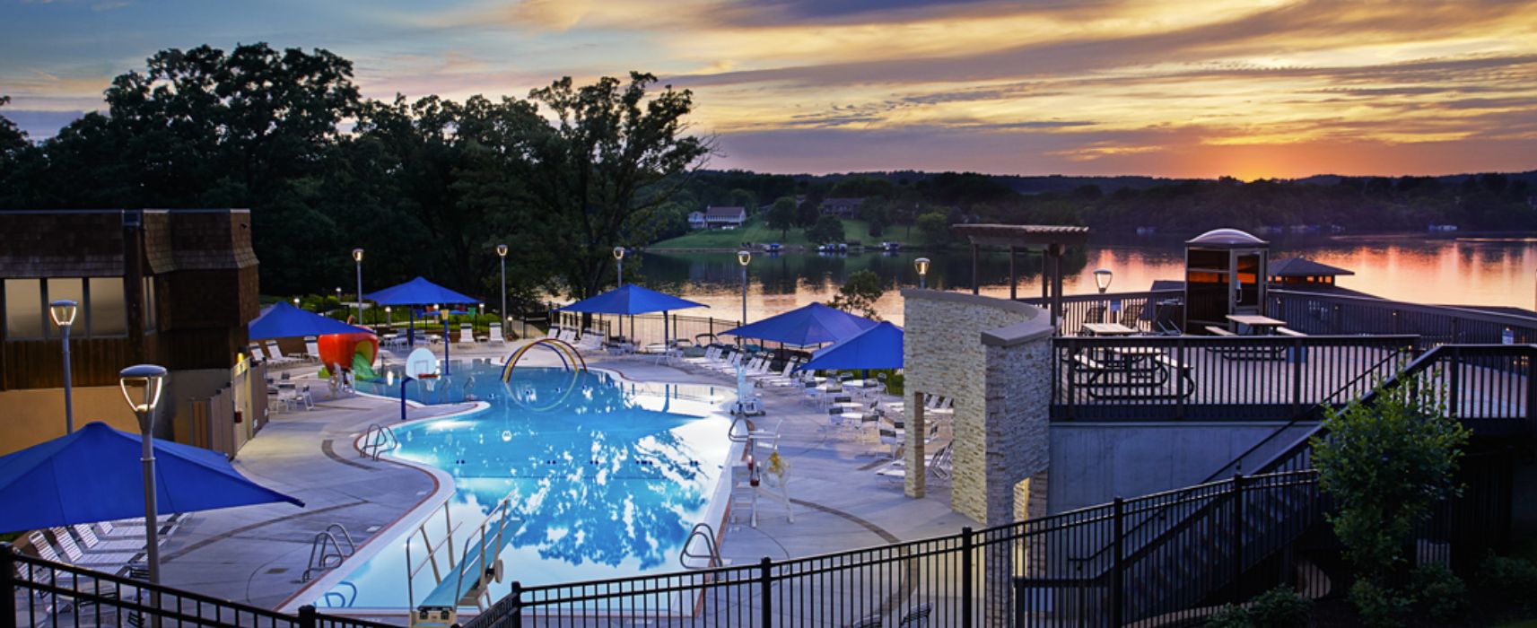 A swimming pool with lounge chairs and umbrellas, surrounded by a stone deck and a view of a lake at sunset.