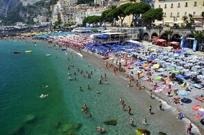 A crowded beach with people swimming and sunbathing, surrounded by colorful umbrellas and beachside cafes.