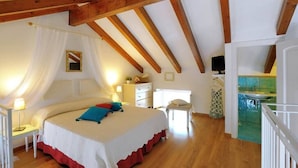 Un dormitorio con techo de madera, cama con ropa de cama blanca y almohadas coloridas, mesita de noche con lámpara y una pequeña silla.