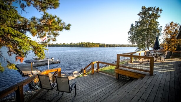 A wooden deck with outdoor seating, overlooking a lake with boats and a forested shoreline.