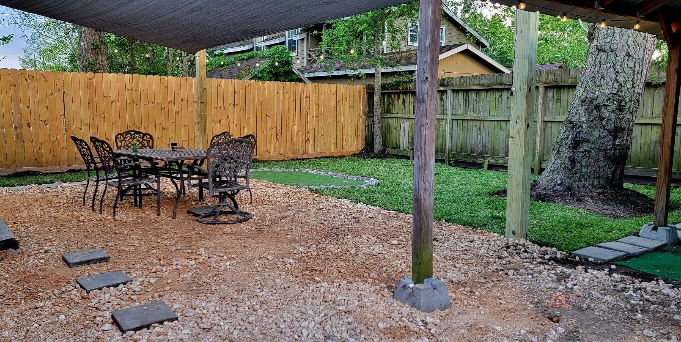 A covered patio area with a table and chairs, surrounded by a wooden fence and a tree.