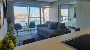 A modern living room with a grey sofa, a small round table, and a view of a marina and buildings through large windows. - Amazing Loft in the Marina (Puerto Vallarta)