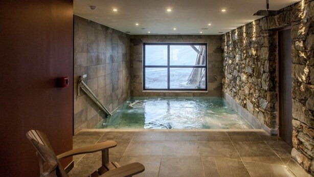 A spa room with a stone wall, a large window, a hot tub, and a wooden chair.