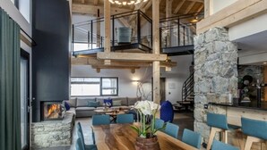 A spacious living area with a wooden dining table, blue chairs, a stone fireplace, and a staircase leading to an upper level.