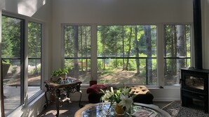 A spacious room with large windows offering a forest view, a round table with a vase of flowers, and a cozy seating area. - Muskoka River Retreat – A Year-Round Paradise (Bracebridge)