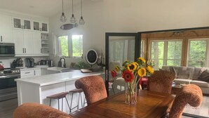 A modern kitchen with white cabinets, a large island, and a dining area with patterned chairs and a wooden table. - Muskoka River Retreat – A Year-Round Paradise (Bracebridge)