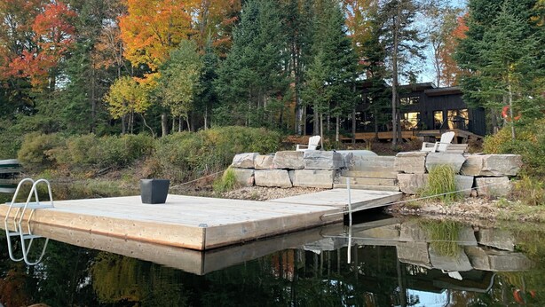 A wooden dock extending into a calm body of water, surrounded by autumn-colored trees and a modern building with large windows. - Muskoka River Retreat – A Year-Round Paradise (Bracebridge)