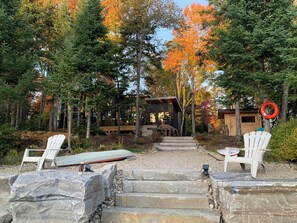 A cabin with a wooden deck, surrounded by trees with autumn foliage. There are two white Adirondack chairs and a canoe.