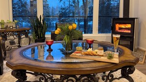 A cozy room with a large window, a fireplace, and a dining table set with snacks and drinks. - Muskoka River Retreat – A Year-Round Paradise (Bracebridge)