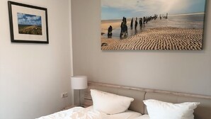 A neatly made double bed with white linens and pillows, a bedside table with a lamp, and a framed picture on the wall.