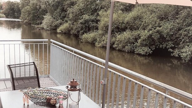 Ein Tisch mit weißer Tischdecke, einem Metallkorb und einem Kerzenständer auf einem Balkon mit Blick auf einen Fluss.