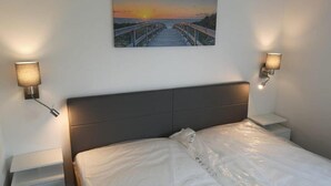 Ein Schlafzimmer mit einem Bett, zwei Nachttischen, einer Wandlampe und einem Gemälde, das einen Sonnenuntergang über einem Steg zeigt.