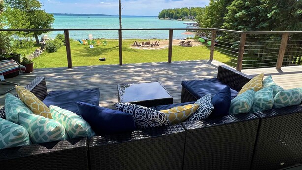 A deck with wicker furniture and cushions, overlooking a grassy area with a view of the sea and trees. - Private Elk Lake Cottage minutes from Elk Rapids (Kewadin)