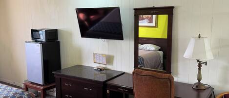 Habitación de hotel con cama, escritorio, silla, televisor, lámpara, espejo y refrigerador.