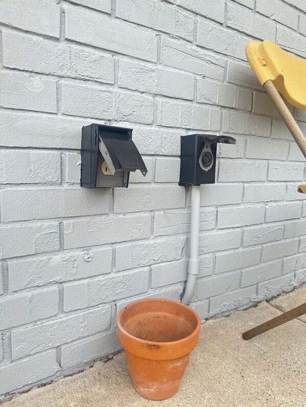 Una pared de ladrillo blanco con dos cajas eléctricas negras y un cubo de terracota en el suelo.