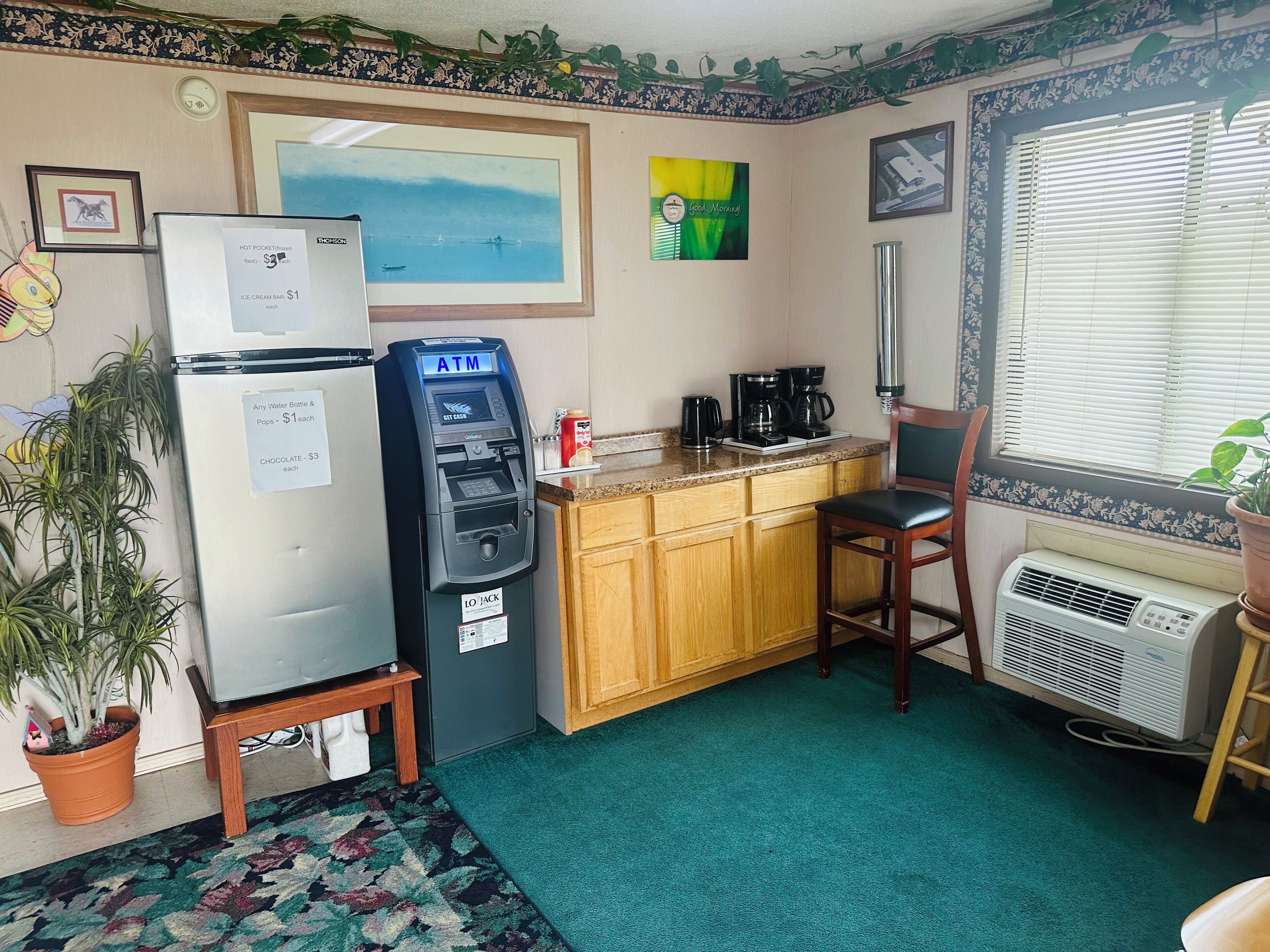A compact kitchen with a refrigerator, an ATM, a coffee maker, and a window with blinds.