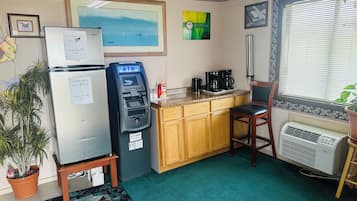 A compact kitchen with a refrigerator, an ATM, a coffee maker, and a window with blinds.