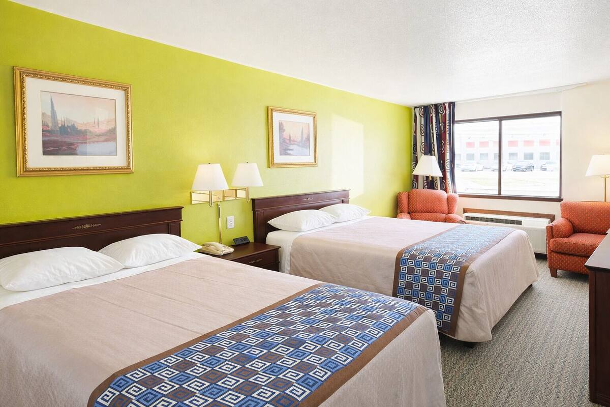 A hotel room with two beds, a chair, a desk, and a window with a view of parked cars.