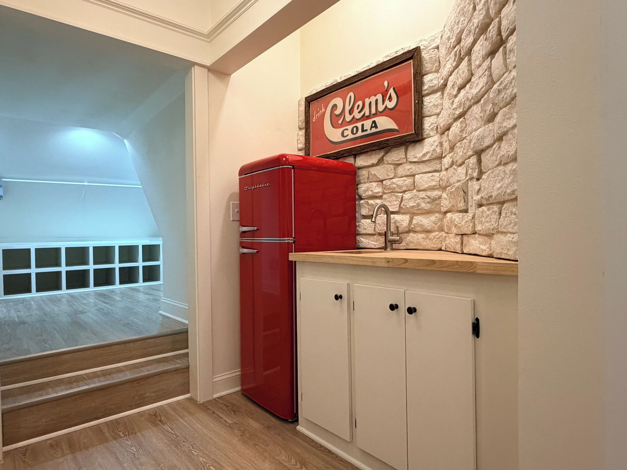 A kitchenette with a red refrigerator, a stone-textured wall, and a sign above the sink that reads 'Clem's Cola.'