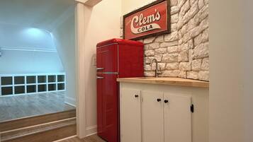 A kitchenette with a red refrigerator, a stone-textured wall, and a sign above the sink that reads 'Clem's Cola.'