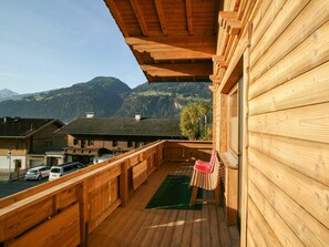 Un balcón de madera con vistas a una cadena montañosa, una alfombra verde y una silla roja.