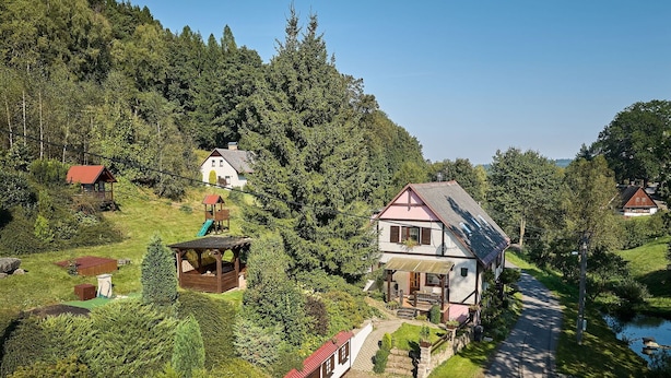 Une grande maison à deux étages, dotée d’un toit en pente, entourée d’une végétation luxuriante et traversée par un petit ruisseau.