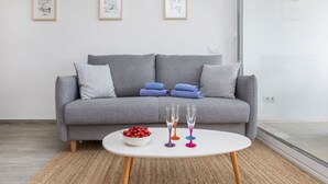 Ein modernes Wohnzimmer mit einem grauen Sofa, zwei blauen Handtüchern, einem weißen Couchtisch mit einer Obstschale und bunten Gläsern, sowie drei gerahmten Kunstwerken an der Wand.