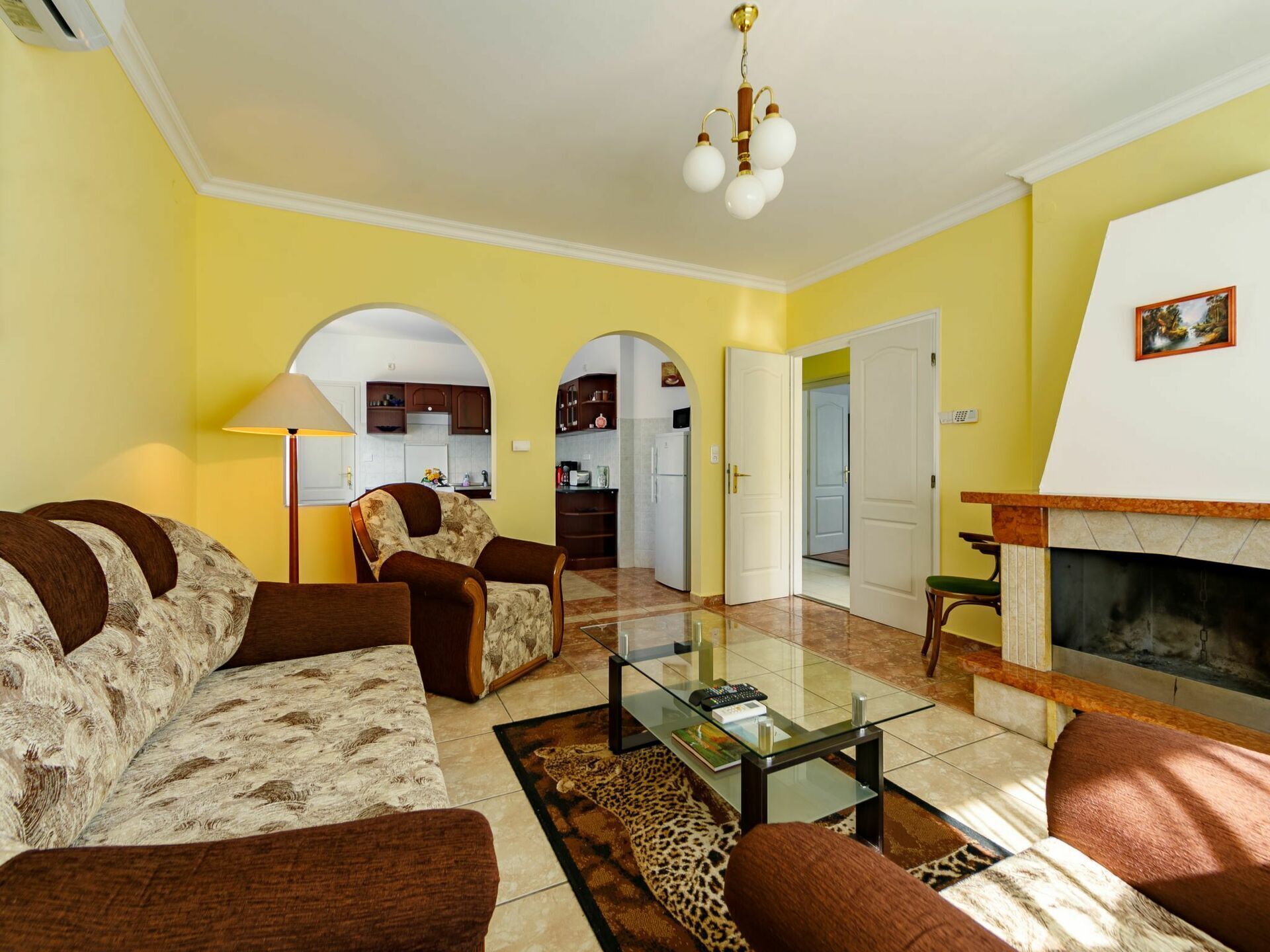 Sala de estar com sofá, poltrona e mesa de centro. Há uma lareira e uma cozinha visíveis por um arco.