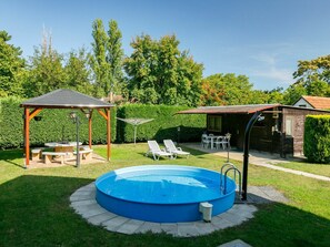 A backyard with a round blue pool, a wooden pergola, and a small wooden building with a red roof.