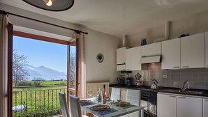 A kitchen with a dining area, a view of the outdoors, and a window with curtains. - Nice apartment for 4 people with WIFI, TV, balcony, pets allowed and panoramic view (Gravedona)