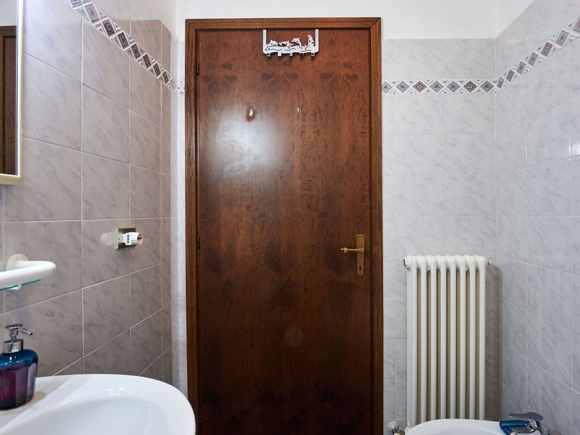 A bathroom with a wooden door, white tiles, a white radiator, and a blue soap dispenser.