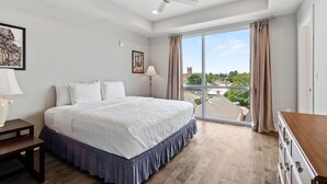A bedroom with a large bed, a ceiling fan, a nightstand, a lamp, and a view of the city through large windows. - Luxury 2 Bed 2 Bath Condo In Bywater with Pool! (New Orleans)