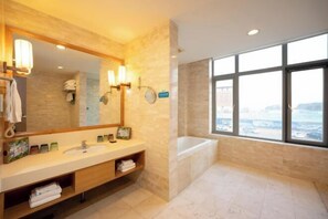 A modern bathroom with a large mirror, a bathtub, and a window offering a city view. - Discoveryland Hotel (Dalian)