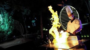 Un chef realiza un espectáculo de fuego en un evento al aire libre, con una estructura metálica circular al fondo.