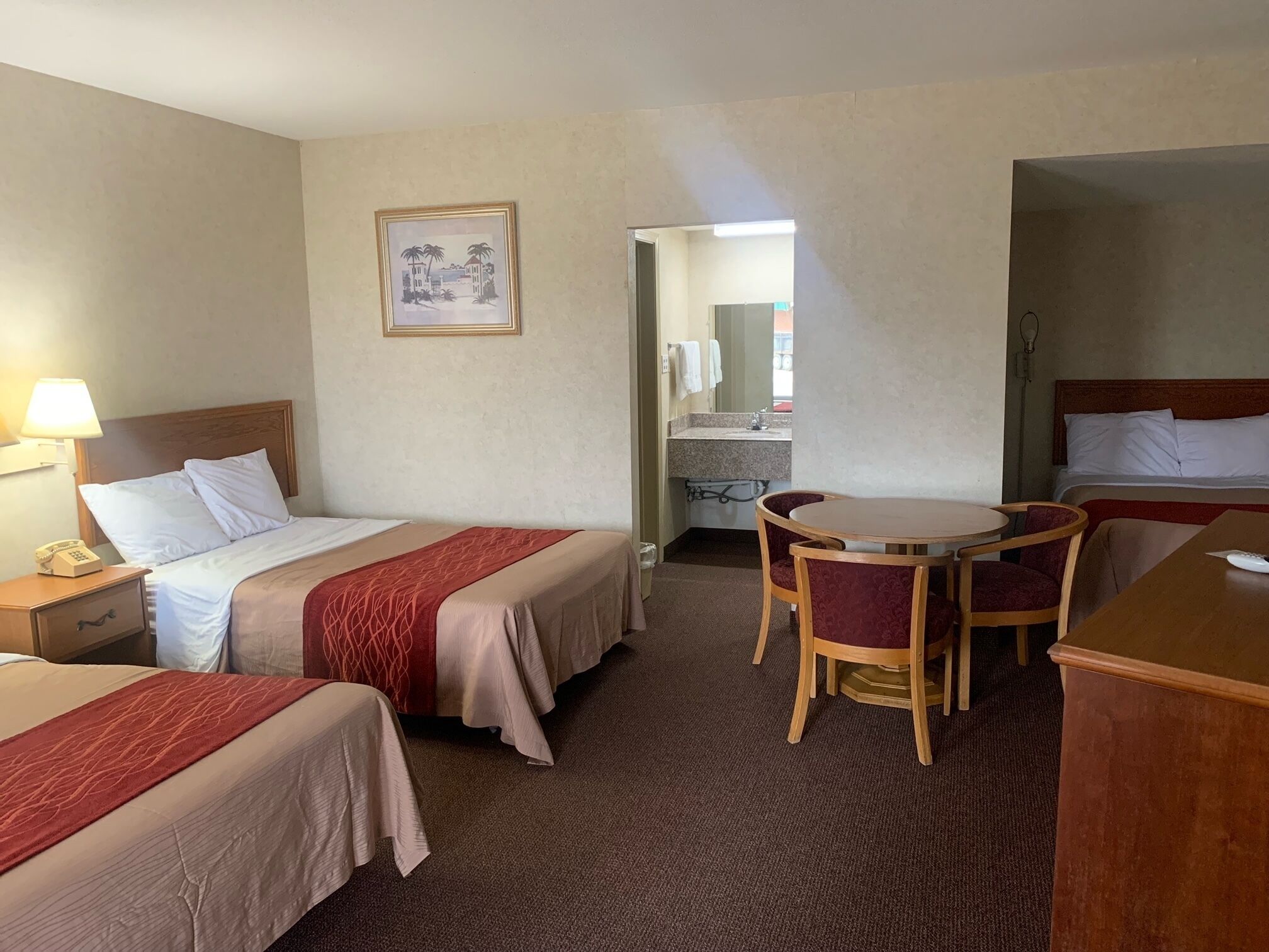 A hotel room with two beds, a desk, a chair, a small table, a lamp, and a painting on the wall.
