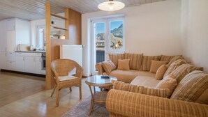 A cozy living area with a sofa, wicker chairs, and a table. A kitchen with white cabinets and a radiator is visible in the background.
