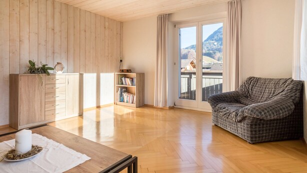 A spacious room with wooden floors, a sofa, a table with a candle, a bookshelf, and a view of mountains through a large window.