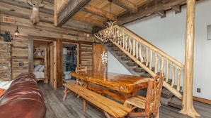 A rustic interior with a wooden dining table, benches, a leather couch, a staircase with wooden railings, and a deer head mounted on the wall.