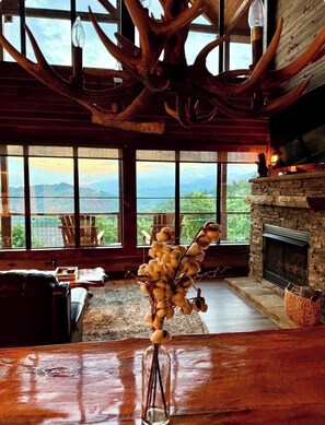 A rustic interior with a large window, a fireplace, and a unique antler chandelier.