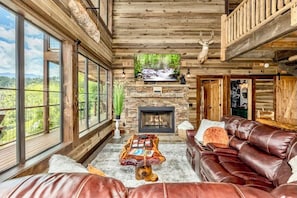 A rustic cabin interior with a stone fireplace, leather sofa, and large windows overlooking a forest.