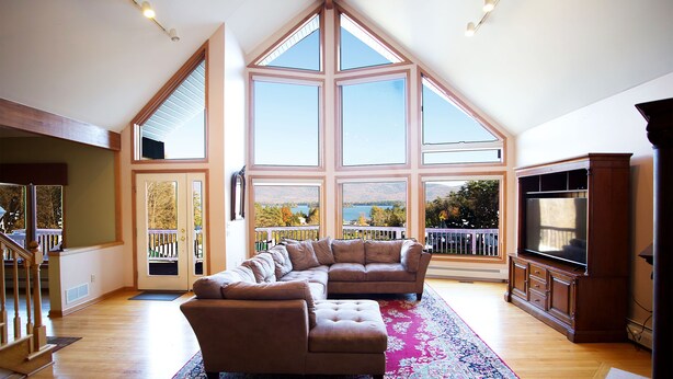 A spacious living room with a large sectional sofa, a flat-screen TV, and a large bay window offering a view of the outdoors. - Lake George House with Beautiful Lake Views and Lake Access 6 Bedrooms 14 People (Lake George)