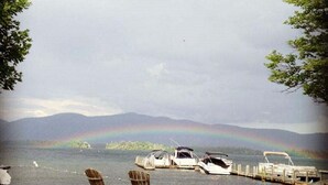 A lakeside view with boats docked, a rainbow, and a grassy area with Adirondack chairs. - Lake George House with Beautiful Lake Views and Lake Access 6 Bedrooms 14 People (Lake George)