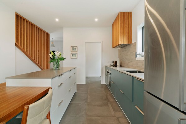 A modern kitchen with wooden cabinetry, a stainless steel refrigerator, and a dining area with a wooden table and chairs. - Jacks Point Family Retreat - Jacks Point Home (Queenstown)