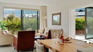 A modern living room with a wooden dining table, leather chairs, and a sofa. There are two wine glasses and a bottle on the table. - Jacks Point Family Retreat - Jacks Point Home (Queenstown)