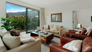 A spacious living room with a large sliding glass door leading to a balcony, a beige sofa, a brown leather sofa, and a glass coffee table with books and decorative items. - Jacks Point Family Retreat - Jacks Point Home (Queenstown)