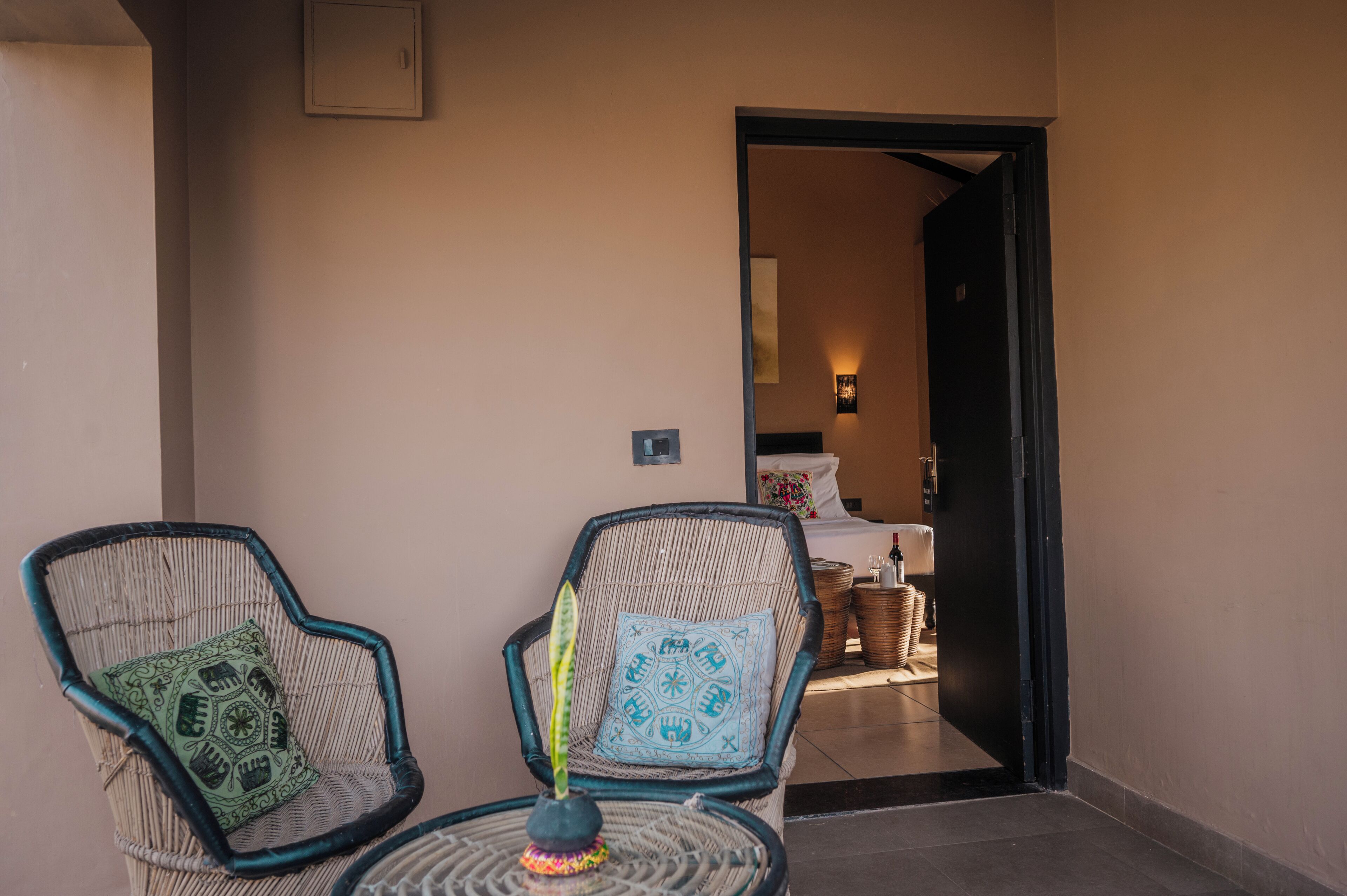 Une pièce comprenant deux fauteuils en osier, une petite table ronde et une porte-fenêtre donnant sur une chambre à coucher avec un lit et une table de chevet.